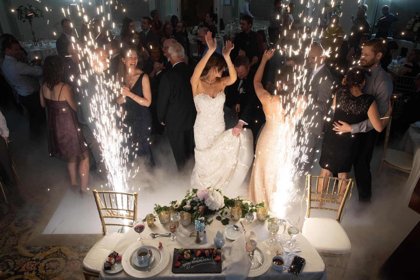 A bride and a bridesmaid dancing on a low-lying cloud effect between cold sparkler fountains, behind a head table set with flowers and dessert