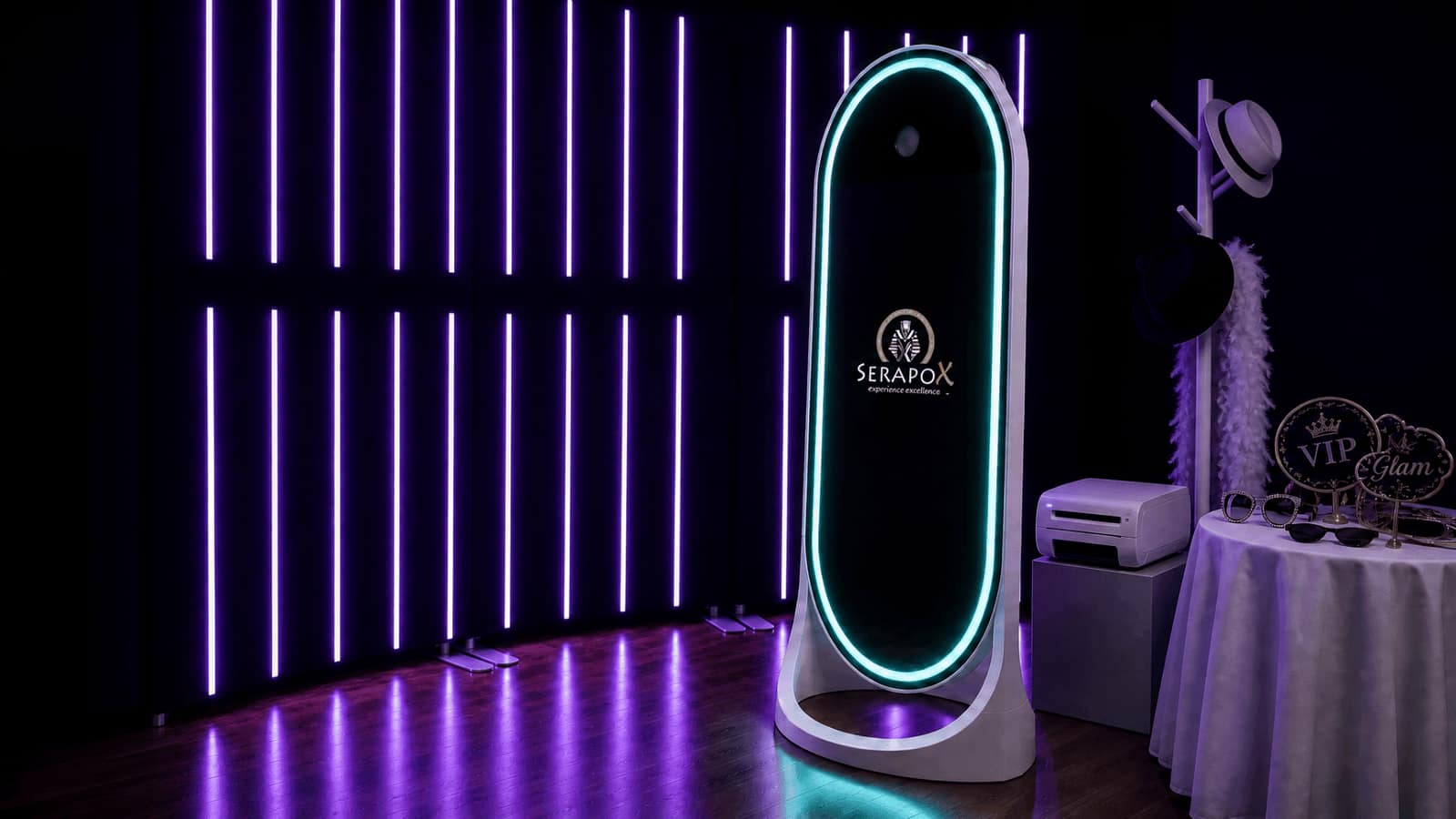 The Mirror Studio: a white oval mirror photo booth with a glowing teal frame and the SerapoX logo, beside a photo printer and a props table, under purple light bars